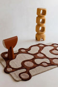 A modern abstract area rug with organic wavy patterns in dark brown and beige, styled with a terracotta-colored sculptural stool and a wooden block tower on a light beige background.