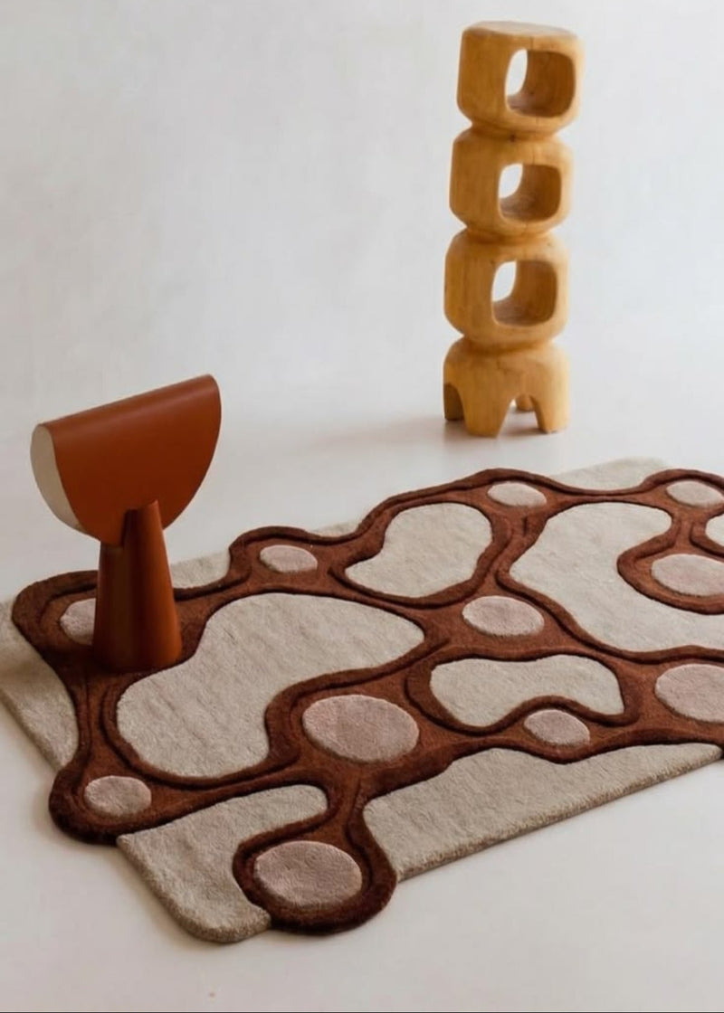 A modern abstract area rug with organic wavy patterns in dark brown and beige, styled with a terracotta-colored sculptural stool and a wooden block tower on a light beige background.