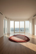 A round area rug with concentric circular patterns in shades of rust red, terracotta, and beige, placed on light wood flooring in a bright, modern room with large windows.