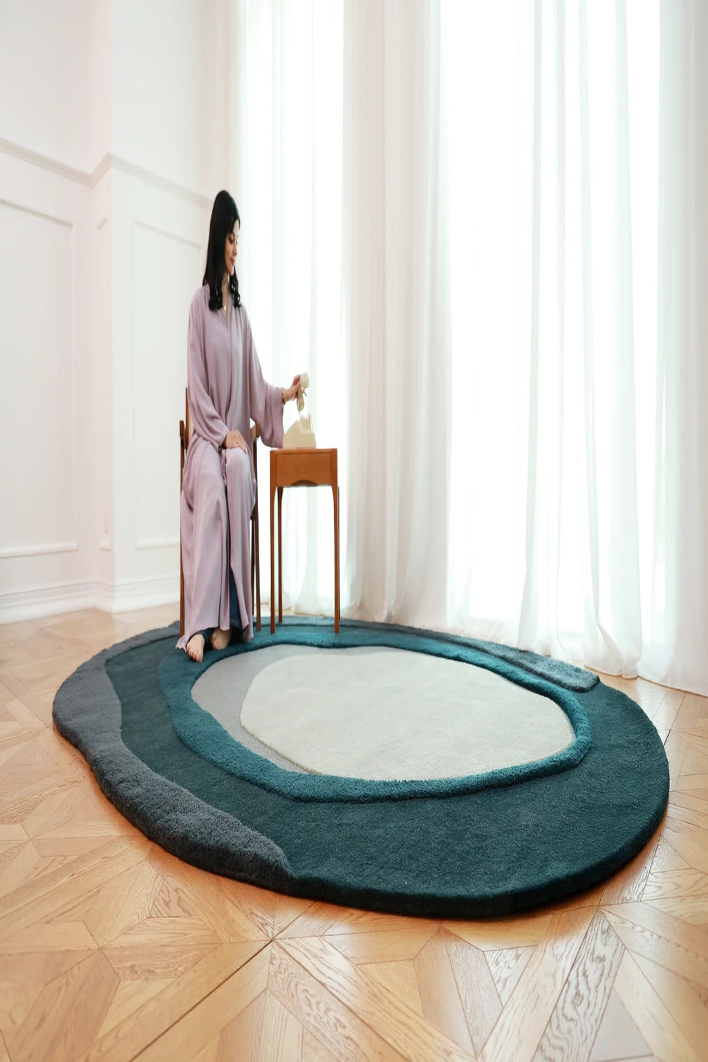 Woman sitting on a chair next to a round rug in a room with large windows.