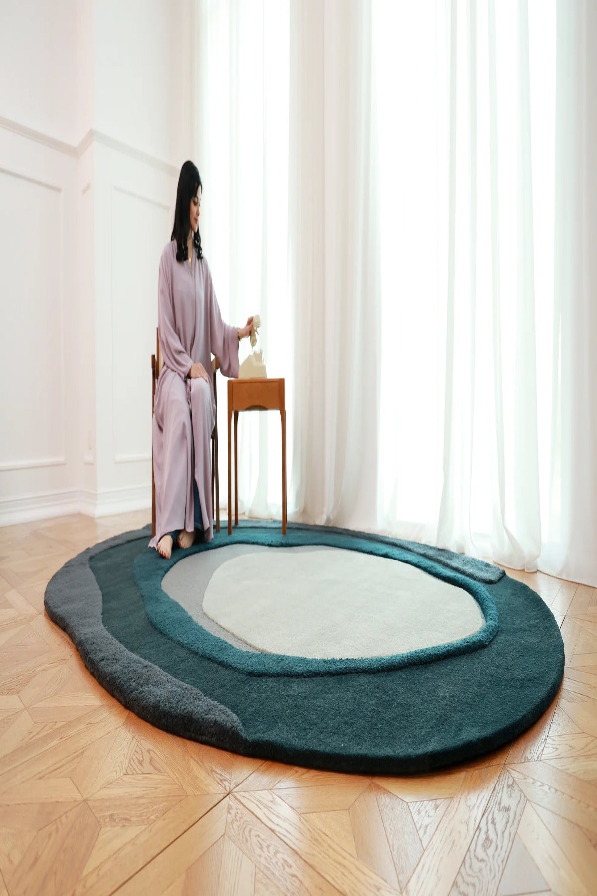 Woman sitting on a chair next to a round rug in a room with large windows.