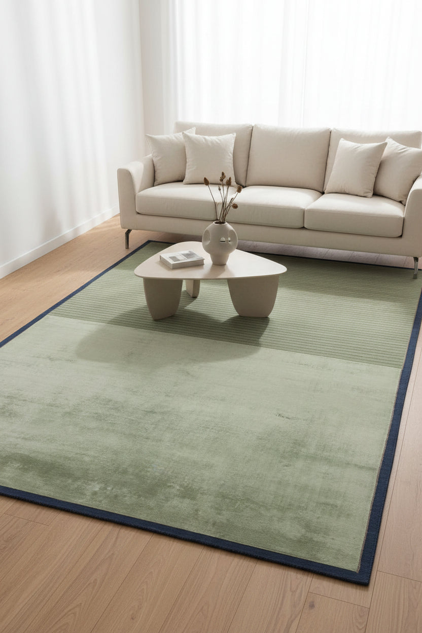 A rectangular area rug with a two-tone design, featuring a textured light sage green upper half and a ribbed olive green lower half, finished with a dark blue border.