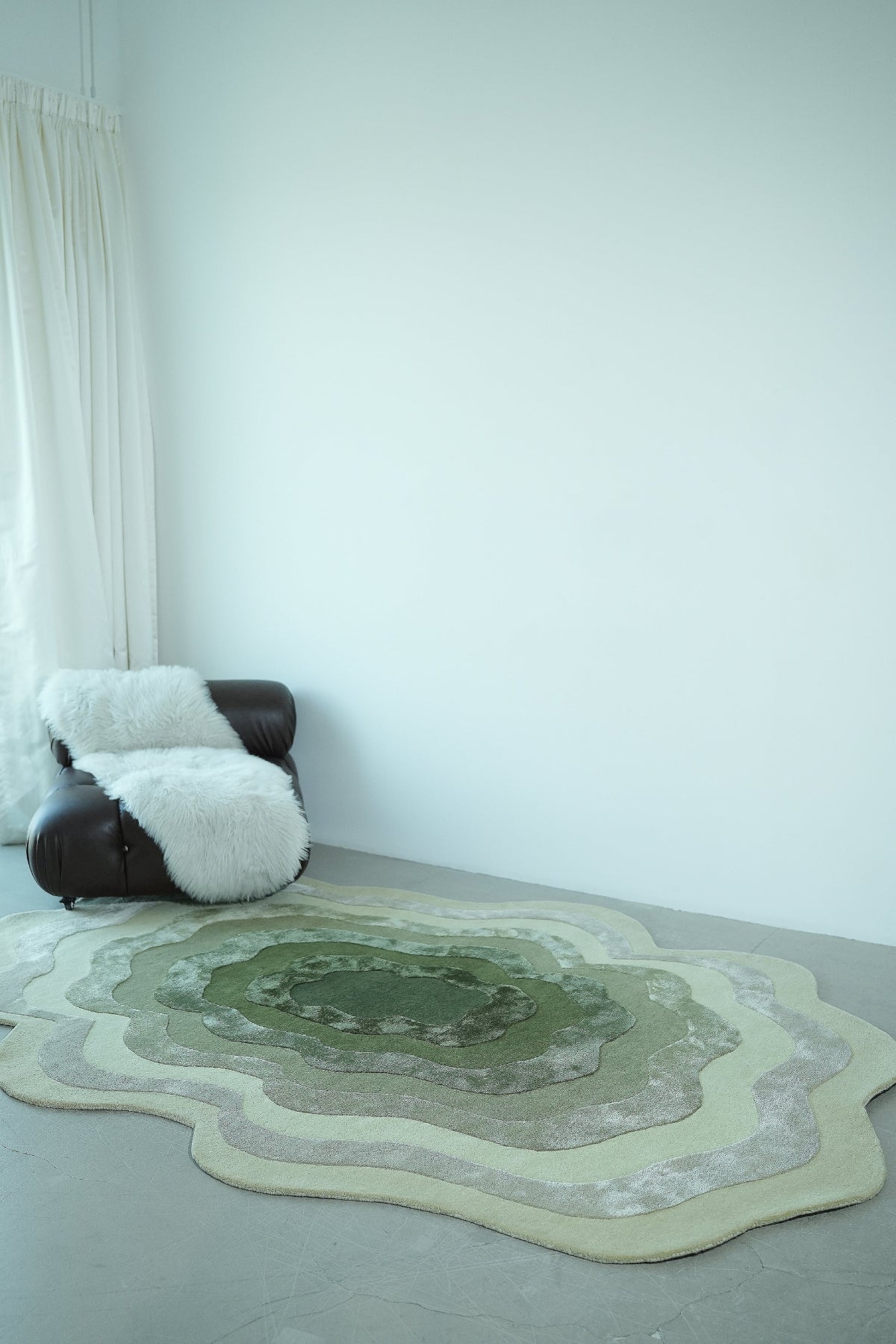 Round green and beige rug on a light gray floor with a white wall and curtain in the background.